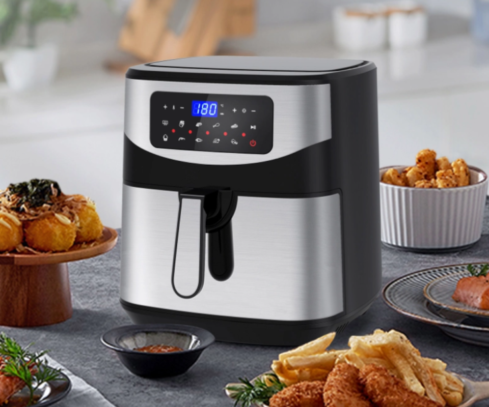 A square stainless steel air fryer sits on a charcoal kitchen bench surrounded by brown fried food. A pale background is behind.