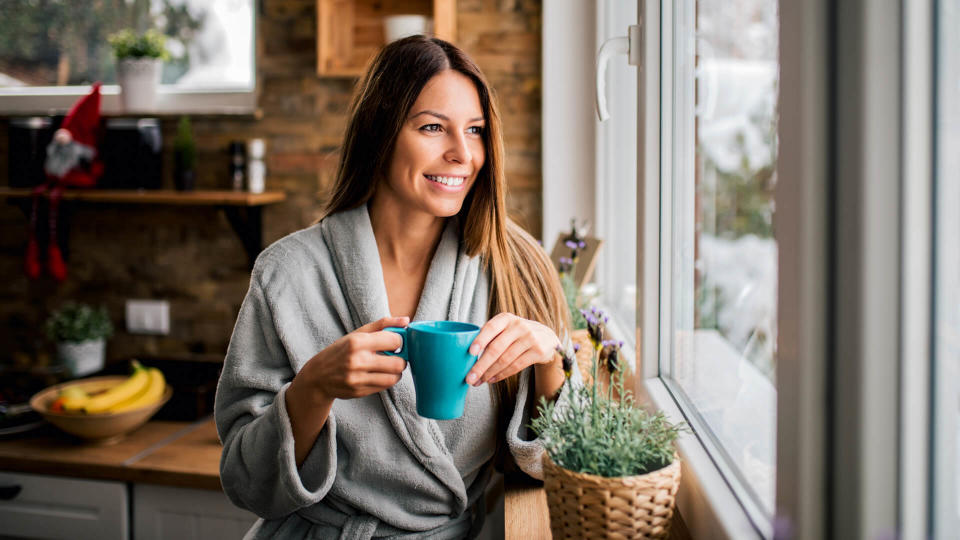 Smiling young woman in bathrobe drinking coffee in the morning, looking through window.