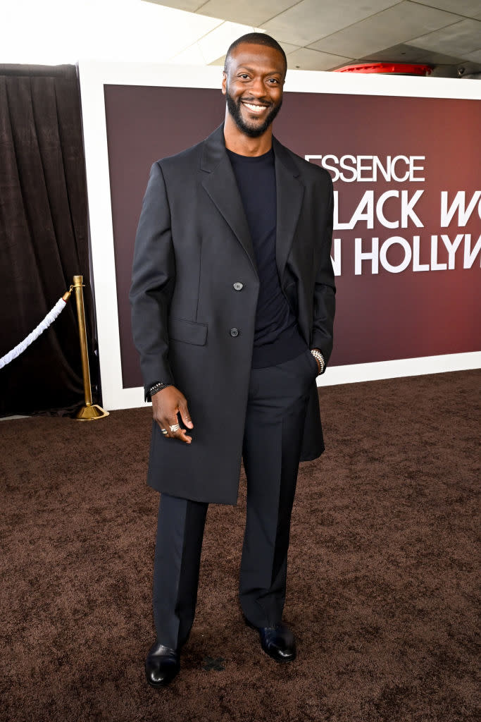 Aldis smiles on the red carpet in a dark suit with long buttoned blazer and dress shoes
