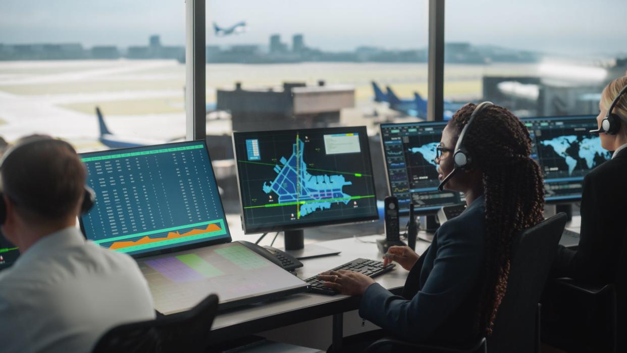 Female Air Traffic Controller with Headset Talk on a Call in Airport Tower. Office Room is Full of Desktop Computer Displays with Navigation Screens, Airplane Flight Radar Data for the Team.