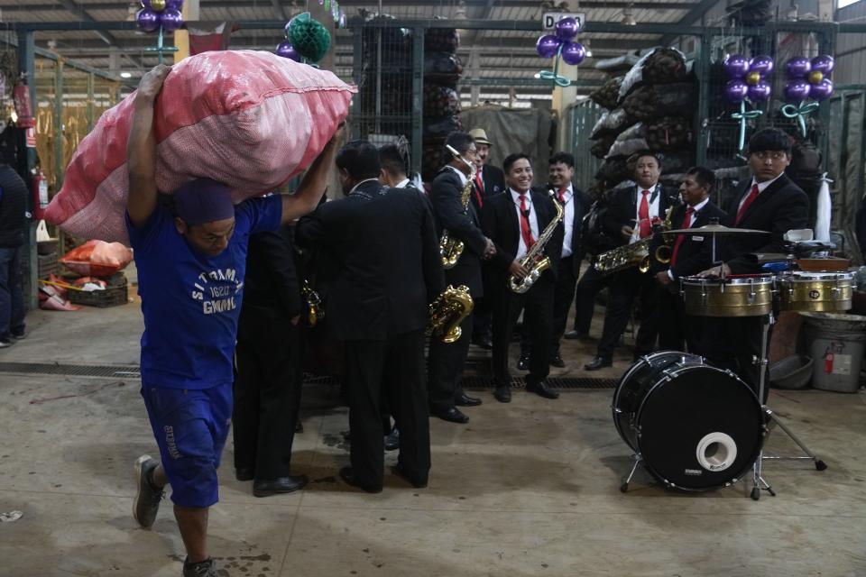 Un cargador lleva a sus espaldas un costal de papas, mientras en el fondo la banda "Los amigos" ofrece un show musical para celebrar el Día Internacional de la Papa en un mercado en las afueras de Lima, Perú, el jueves 30 de mayo de 2024. (AP Foto/Martín Mejía)