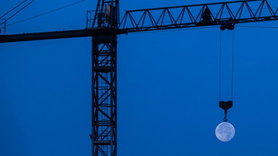 the sky appears a dark blue hue while a silhouette of a crane appears to hook the moon which shines in the background.