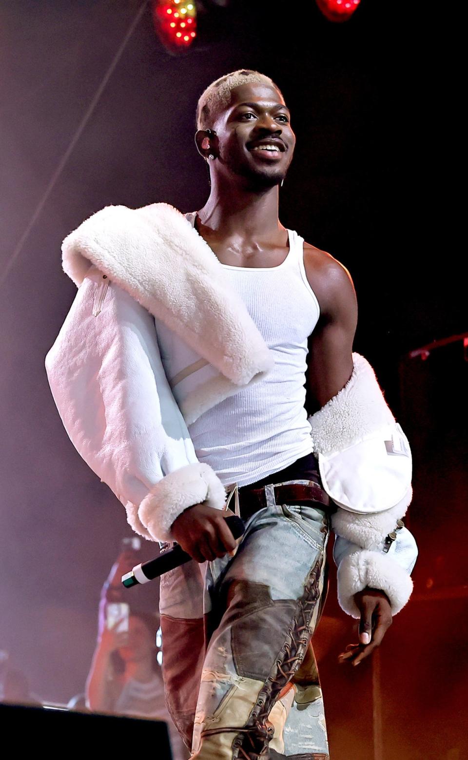 Lil Nas X is performing on stage, wearing a tank top, a fur coat draped over his shoulder, and patchwork jeans