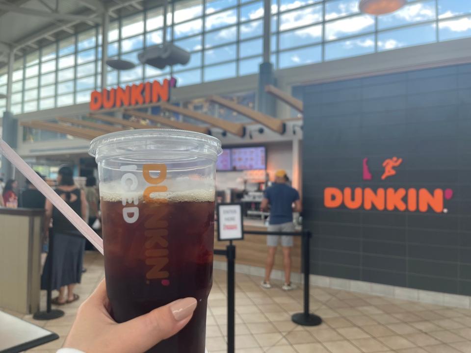 A hand holding a cup of iced coffee from Dunkin' in front of a rest stop.