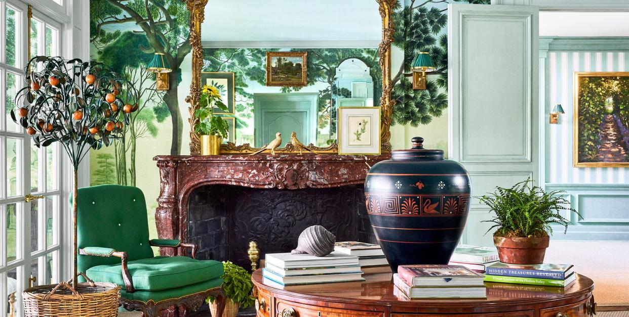 an entryway has a fireplace with a decorative marble surround and ornate mirror above, wallpaper with floor to ceiling trees, a round polished wood pedestal table with a vase and books, and a green cushioned armchair