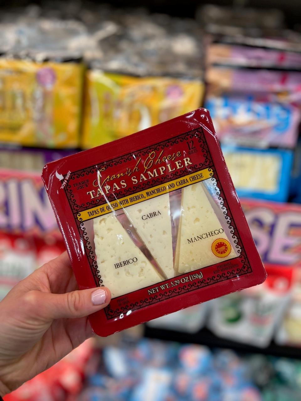 Hand holding a 'Tapas Sampler' cheese package with Iberico, Cabra, and Manchego cheeses at a grocery store