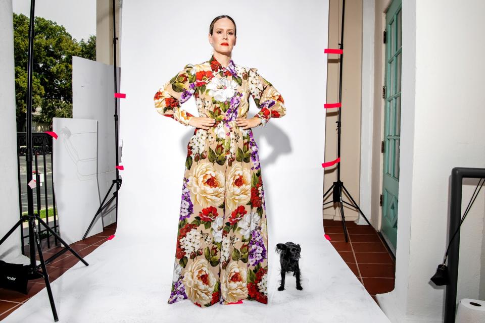 A woman in a dress stands on a white backdrop with her dog.