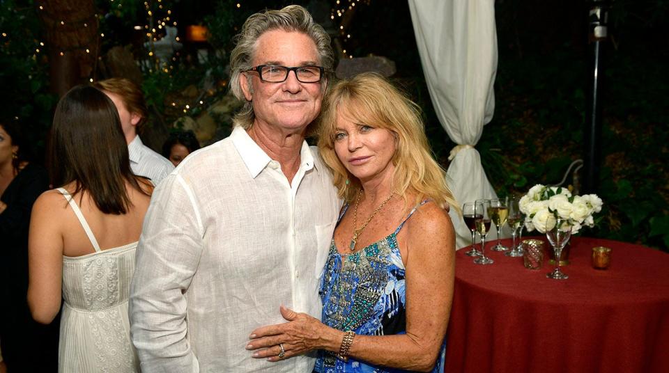 Kurt Russell in a white shirt and Goldie Hawn in a blue printed dress pose for a photo