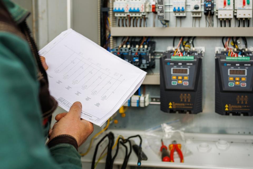 An electrician reads a document with diagrams while assessing an electrical system in a wall.