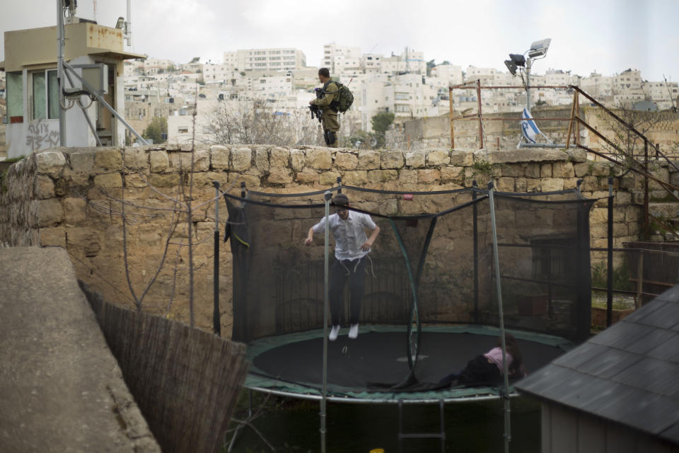 FILE - In this March 7, 2019, file photo, settlers jump on a trampoline as an Israeli solider stands guard in the Israeli controlled part of the West Bank city of Hebron. Israel's premier human rights group has begun describing both Israel and its control of the Palestinian territories as a single "apartheid" regime, using an explosive term that the Israeli government and its supporters vehemently reject. (AP Photo/Ariel Schalit, File)