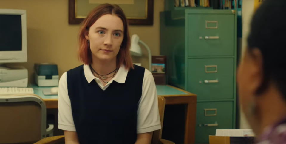 Saoirse Ronan sits at a desk wearing a white shirt and dark vest, talking to an unseen person in a room with office furniture in the background