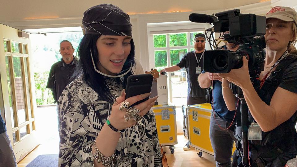 Billie Eilish, a white teenage girl, stands in a room looking at her phone. She wears a bandana in her hair and has many silver rings. A woman holding a movie camera shoots her.