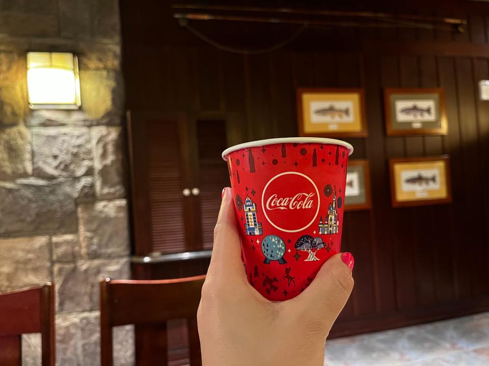 A hand with pink nails holds a red Coca-Cola cup in front of a wooden door