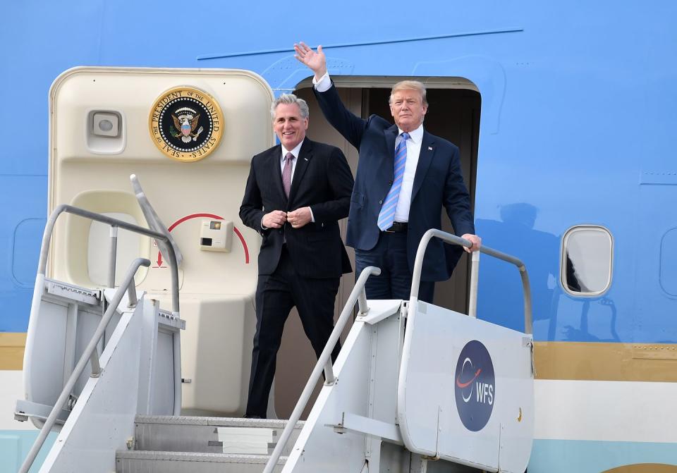 kevin mccarthy and donald trump walk out of an airplane door with a presidential seal on the inside of the door, mccarthy smiles and wears a suit and red tie, trump wears a navy suit with a blue tie and waves