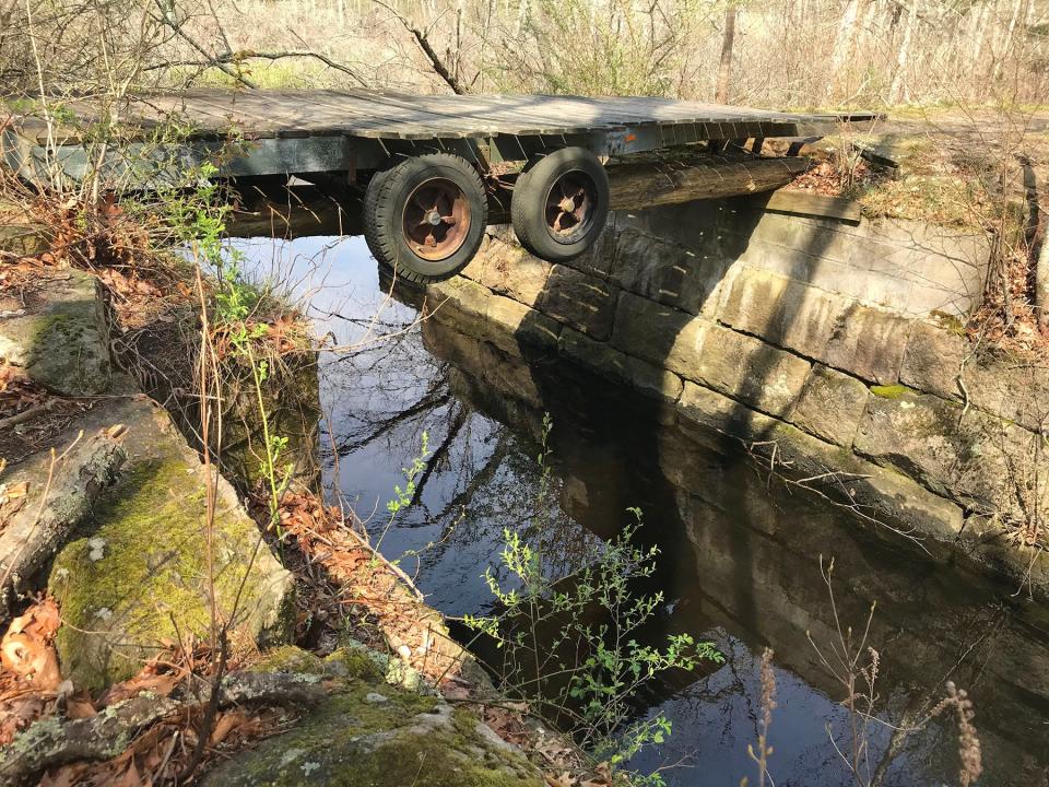 A bridge built from a flat wooden trailer, with wheels still attached, crosses a tributary of the Wood River.