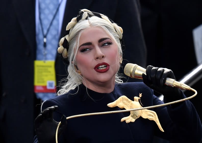 Lady Gaga singing into a golden microphone, wearing a black shirt and gold bird pendant