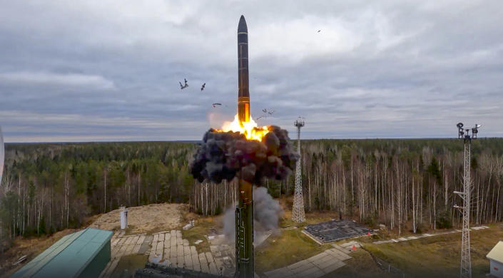 Missile rising from smoke and flames moments after takeoff near a green building and towers in a clearing of trees against a clouded sky.