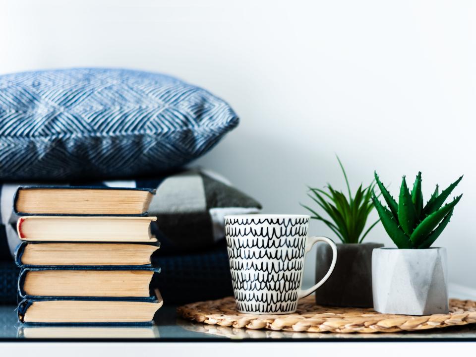 Cozy home interior decor: white and black cup, stack of books, plants in pots on a wicker