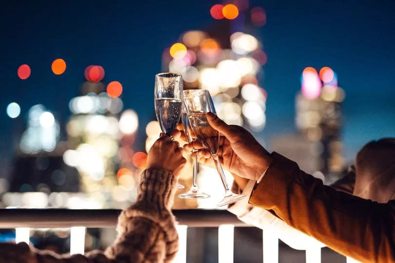 Group of multiracial businesspeople having Friday night drinks at rooftop in office building. Drinking white wine, champagne and prosecco against London city scape. Multiracial business team discussing work and life after work at night, in urban balcony overlooking city, Liverpool, London. Holding wine glasses.