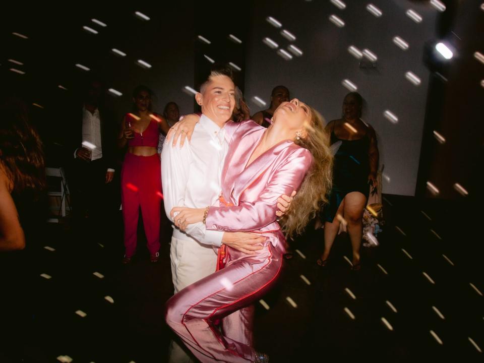 A bride in a white suit and a bride in a pink, shiny jumpsuit dance.