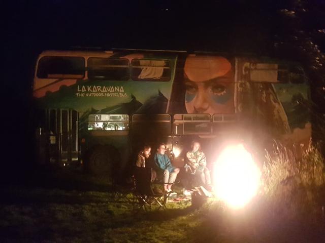 a group of people around a campfire with the double decker bus in the background