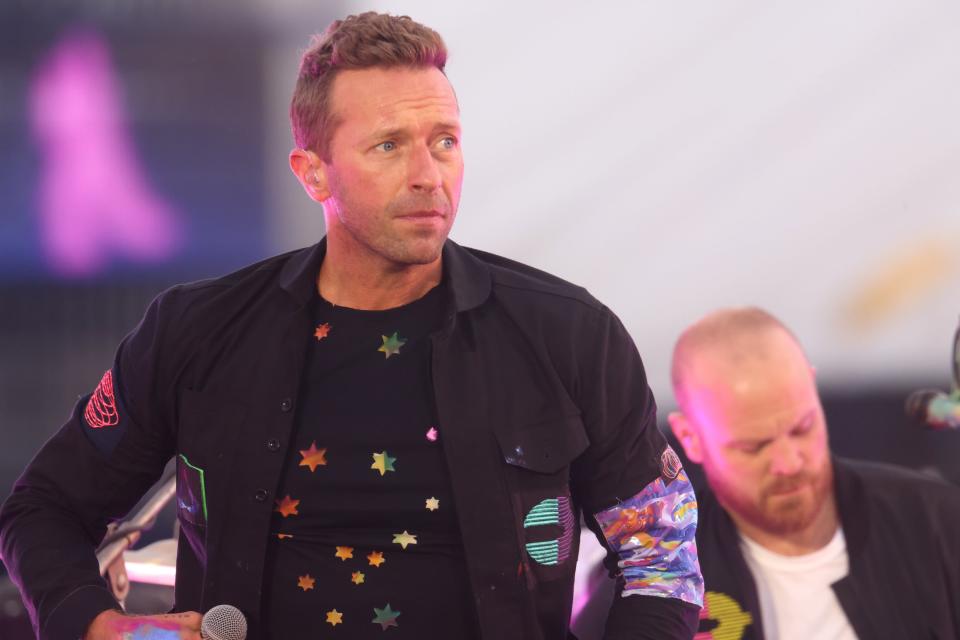 Chris Martin performing on stage with colorful star designs on his black shirt, microphone in hand