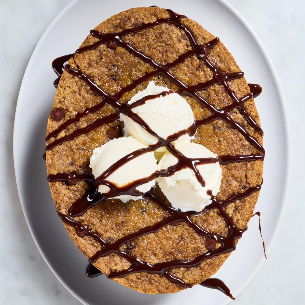 large chocolate chip cookie topped with vanilla ice cream and drizzled with chocolate sauce on top of a white platter