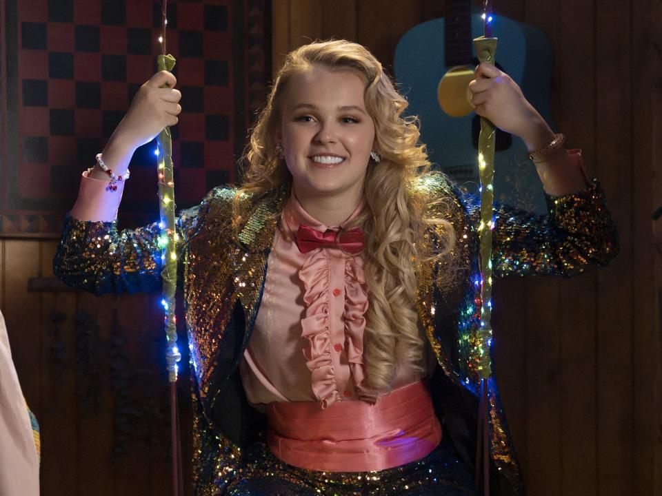 jojo siwa on high school musical the musical the series, wearing a pink shirt with a ruffled button line, a red bowtie, and a sparkling rainbow jacket, sitting on a swing lit with sparkling, multicolored lights