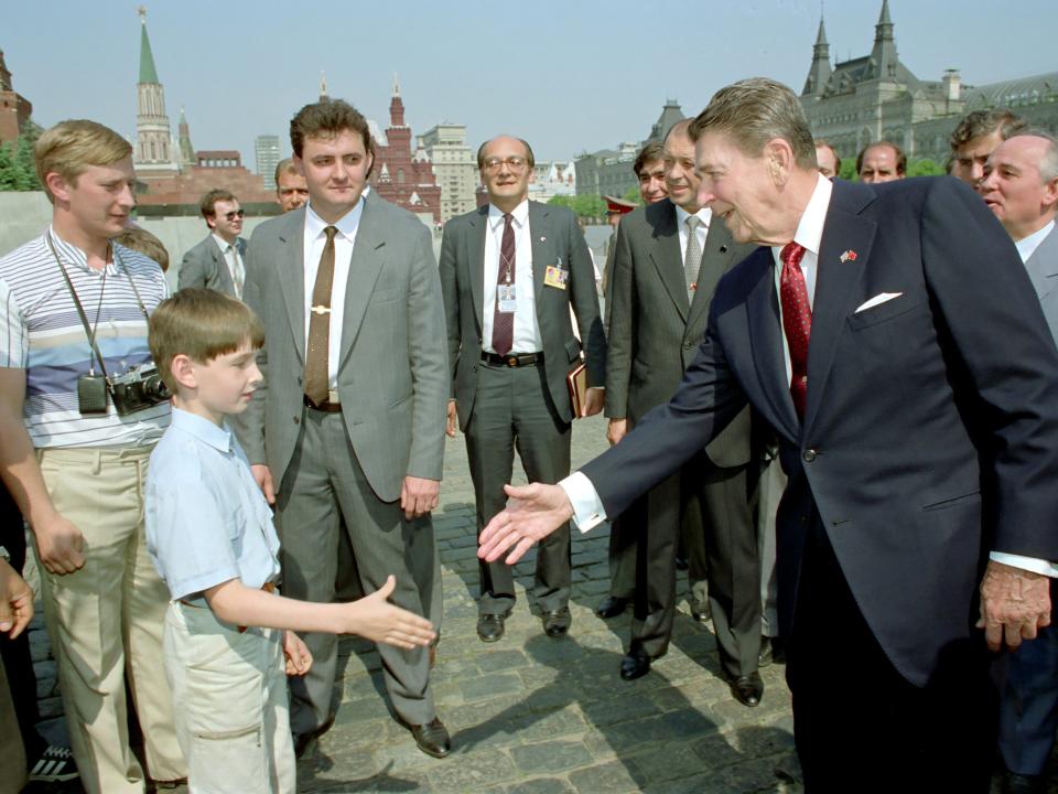 Ronald Reagan le da la mano a un niño mientras Mikhail Gorbachev (der.) observa durante un recorrido por la Plaza Roja de Moscú. ¿Podría también mostrar a un joven Vladimir Putin (izq.) espiando al presidente de Estados Unidos? (Pete Souza/White House via Getty Images)