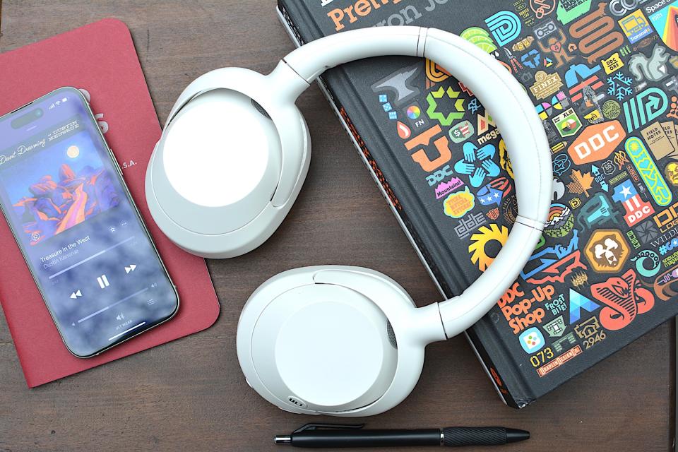 <p>White headphones sitting on a wooden table from above with a red notebook and phone on the left and a black book with logos on the cover on the right/underneath.</p>
