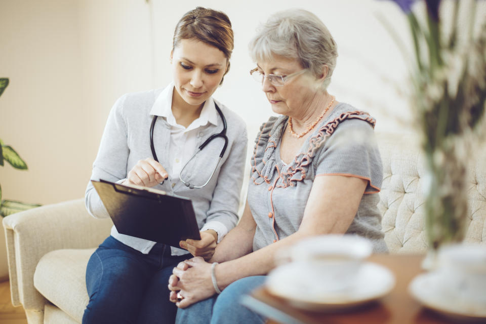 Latin woman wearing a lab coat over blue surgical scrubs is speaking with an elderly woman in a wheelchair. As she speaks, she is smiling and touching the older woman on the shoulder.