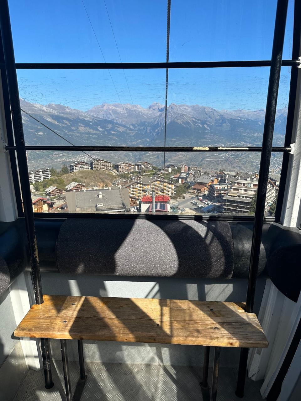 inside of a gondola, a wooden bench beneath a glass wall with a small swiss town in the mountains