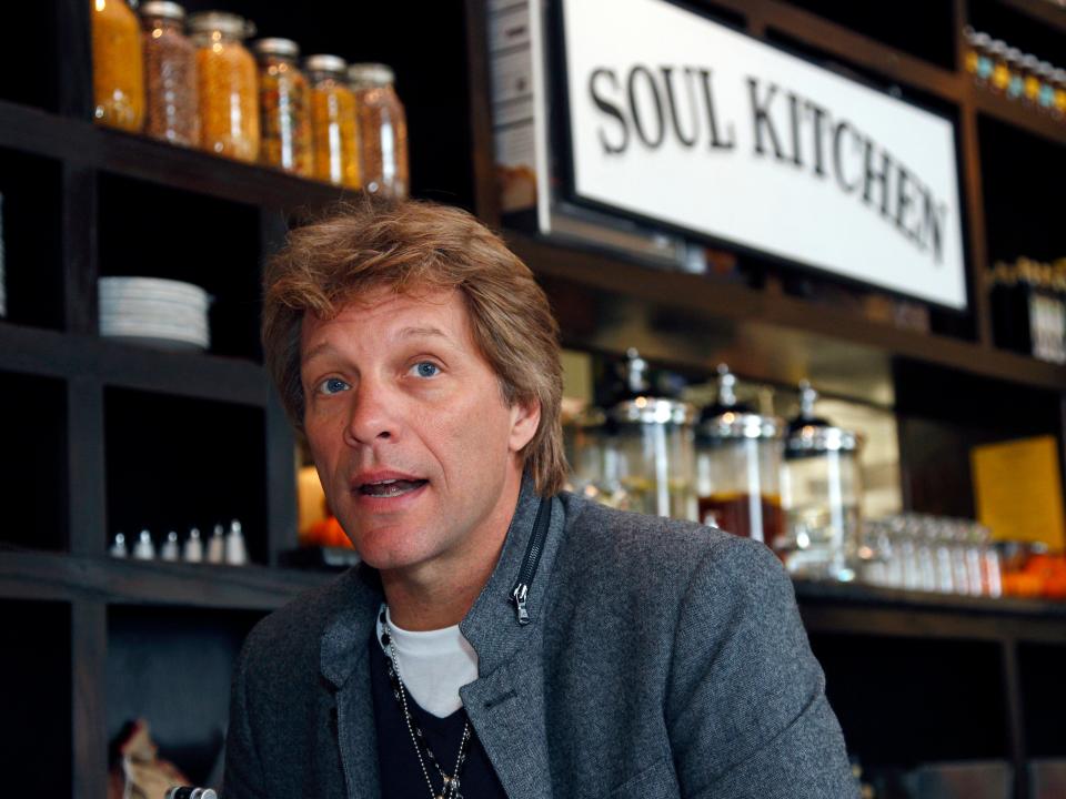Closeup of Bon Jovi talking and sitting in front of a Soul Kitchen sign and shelves with ingredients on display.