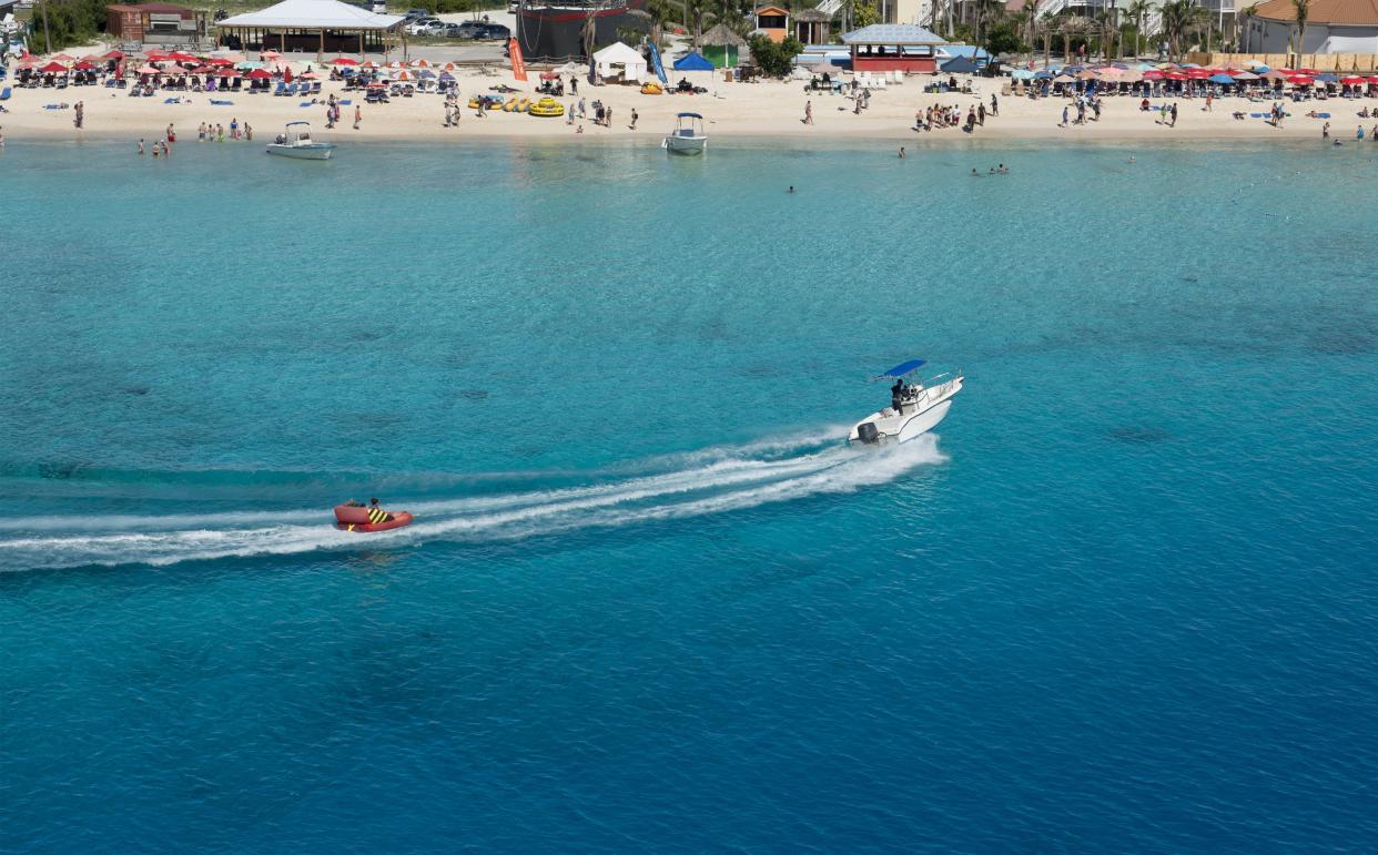 Motorboat pulling an inflatable seat over the turquoise waters of the Caribbean Sea in front of a busy beach. Grand Turk Island, Turks and Caicos Islands.