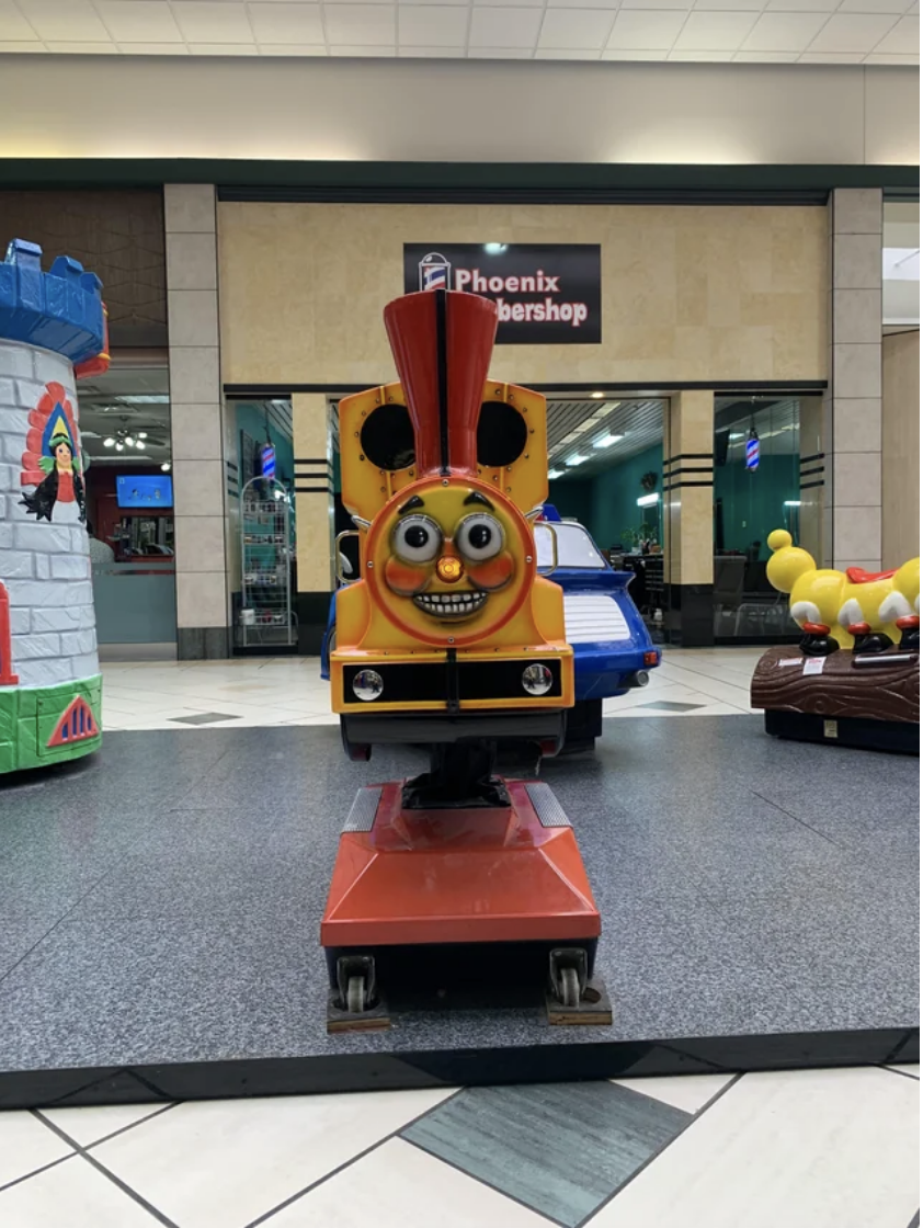 A small children's stationary train ride in a local mall that has big, wide eyes, defined rosy cheeks, and a huge, scary smile