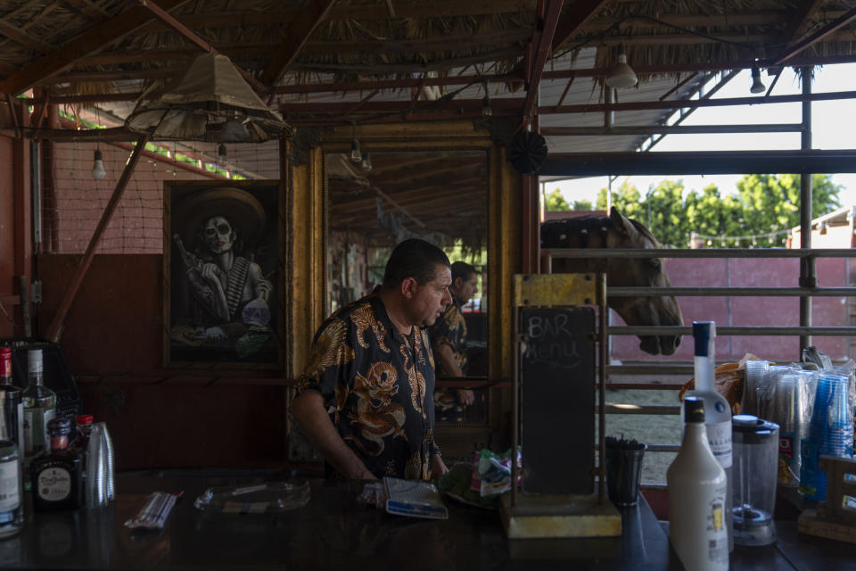 A horse rests in a stable as bartender Graciano Marin readies a bar ahead of a baptism ceremony at Rancho El Refugio date palm ranch owned by Carlos Ulloa in Twentynine Palms, Calif., Saturday, June 15, 2024. (AP Photo/Jae C. Hong)