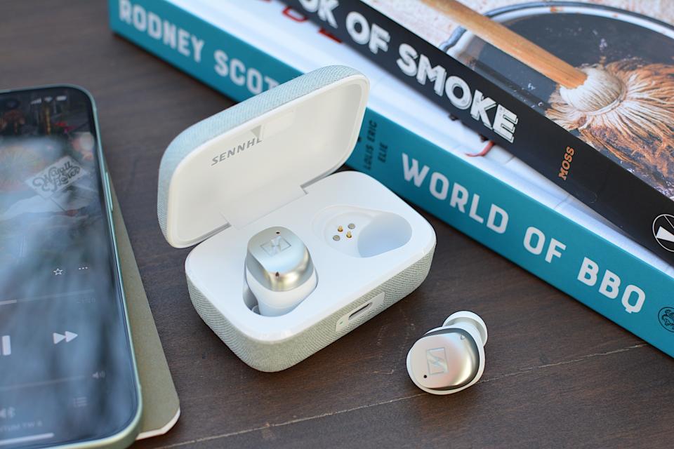 The white/silver Sennheiser Momentum True Wireless 4 headphones sit in their case with the lid open, one earbud sitting on its side on a wooden table.