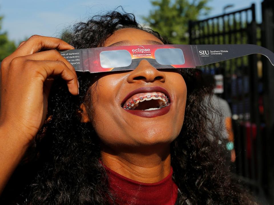 A cheerleader smiles as she looks up at the sky holding eclipse glasses.