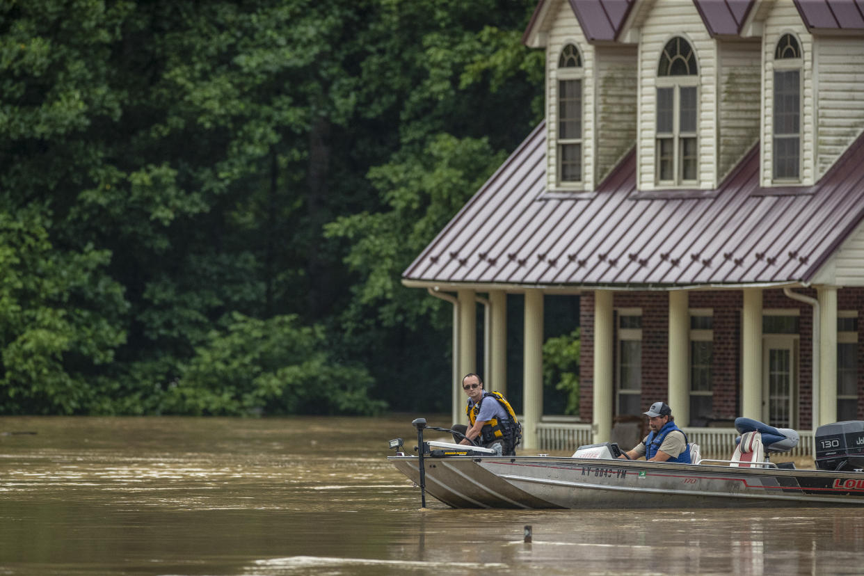 Two men in life jackets navigate past a flooded home in a boat.