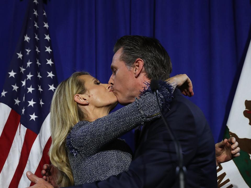 Jennifer Siebel Newsom and Gavin Newsom kiss in front of an American flag.