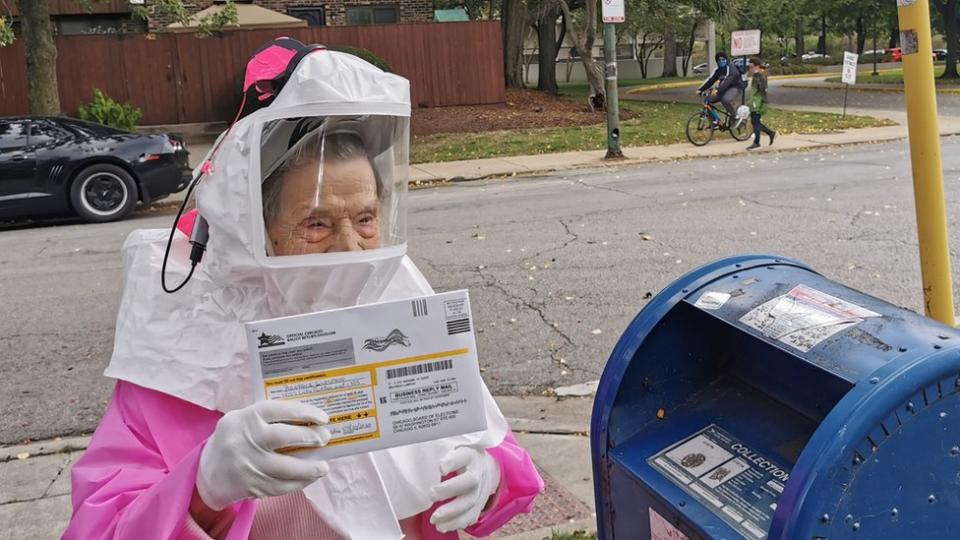 An elderly woman in a protective hood prepared to post her ballot in a mail box