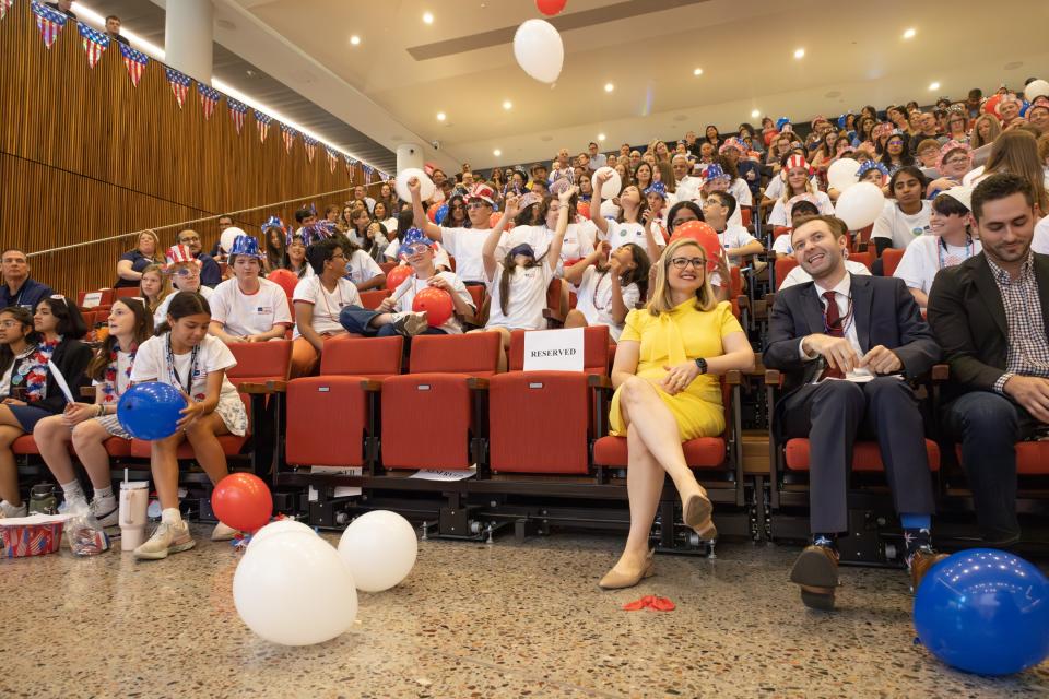 Phoenix Mayor Kate Gallego, dressed in yellow, sits in an auditorium surrounded by young students and balloons.