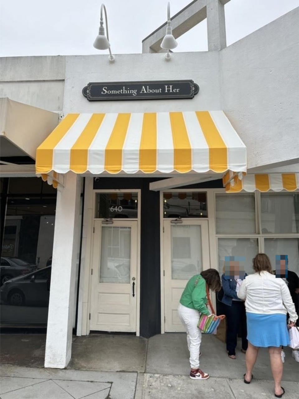 People are standing outside a shop that has a striped awning and a sign reading "Something About Her."