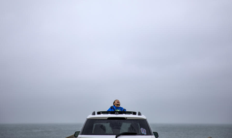 Mason Lambert, 5, looks up at the rain as he leaves the house with his grandmother Karen Lambert, in car at left, and mother Emilie Lambert, right, for his daily outing to one of the few public parking areas still open by the sea during the coronavirus shutdown, April 24, 2020, in Narragansett, R.I. (AP Photo/David Goldman)