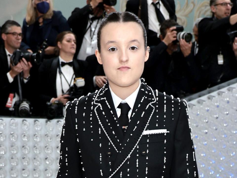 Bella Ramsey standing in front of a group of photographers at the 2023 Met Gala while wearing a black suit and tie with bright silver lining.