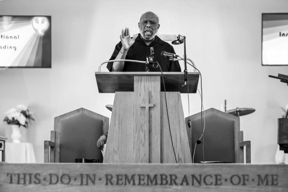 A man stands at a pulpit, speaking with one hand raised.