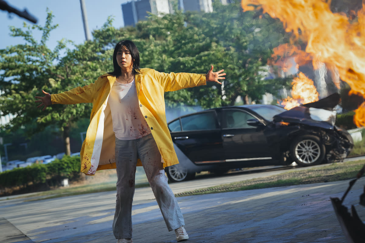  A Korean woman in a yellow coat and bloodstained clothes stands with flaming crashed cars behind her. . 