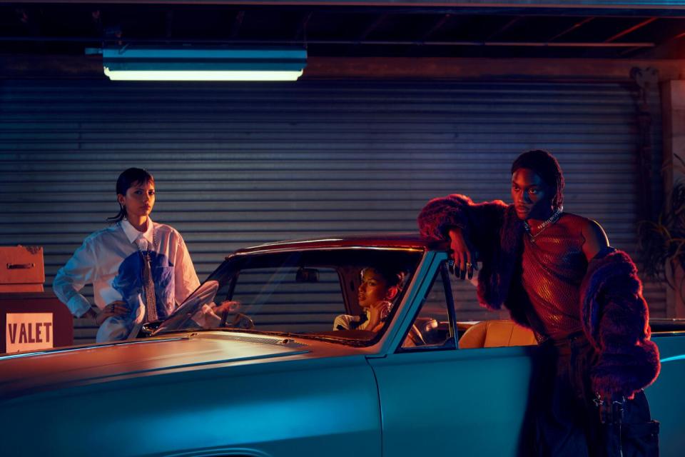 A woman in a car at night adjusts her earring. Two figures lean against the car.