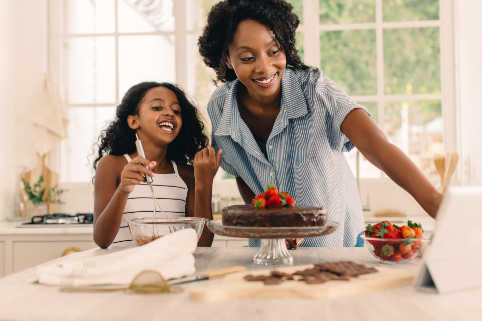 Smiling girl with mother looking at digital tablet while preparing cake in kitchen. Mother and daughter following online recipe while baking cake at home.