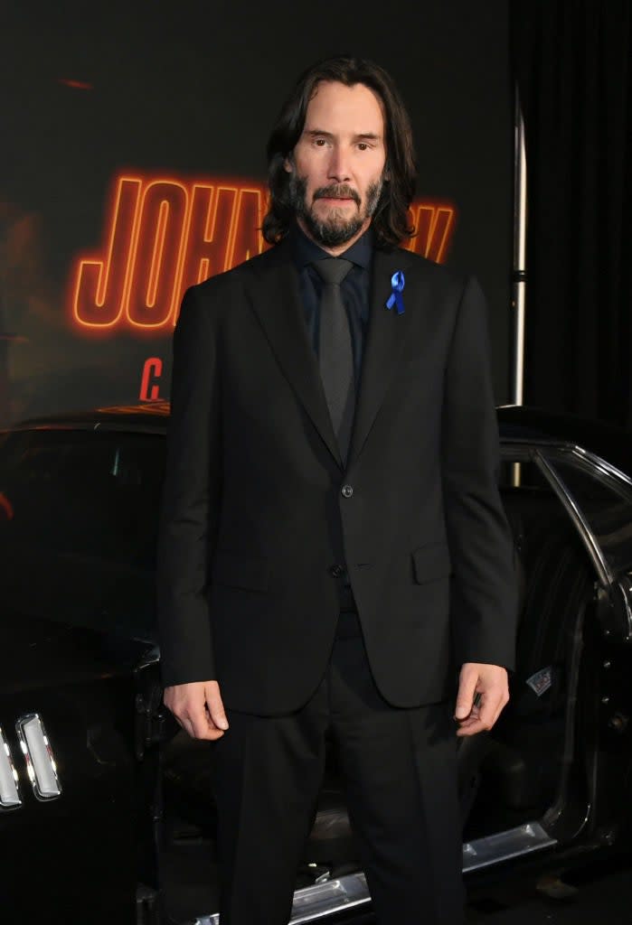 Keanu Reeves in a sharp suit with a ribbon pin, standing in front of a 'John Wick' backdrop